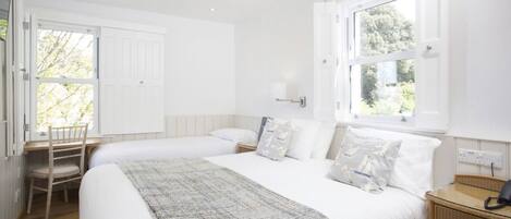 A bedroom with two beds, a desk, a chair, a nightstand, and a window with a view of greenery.
