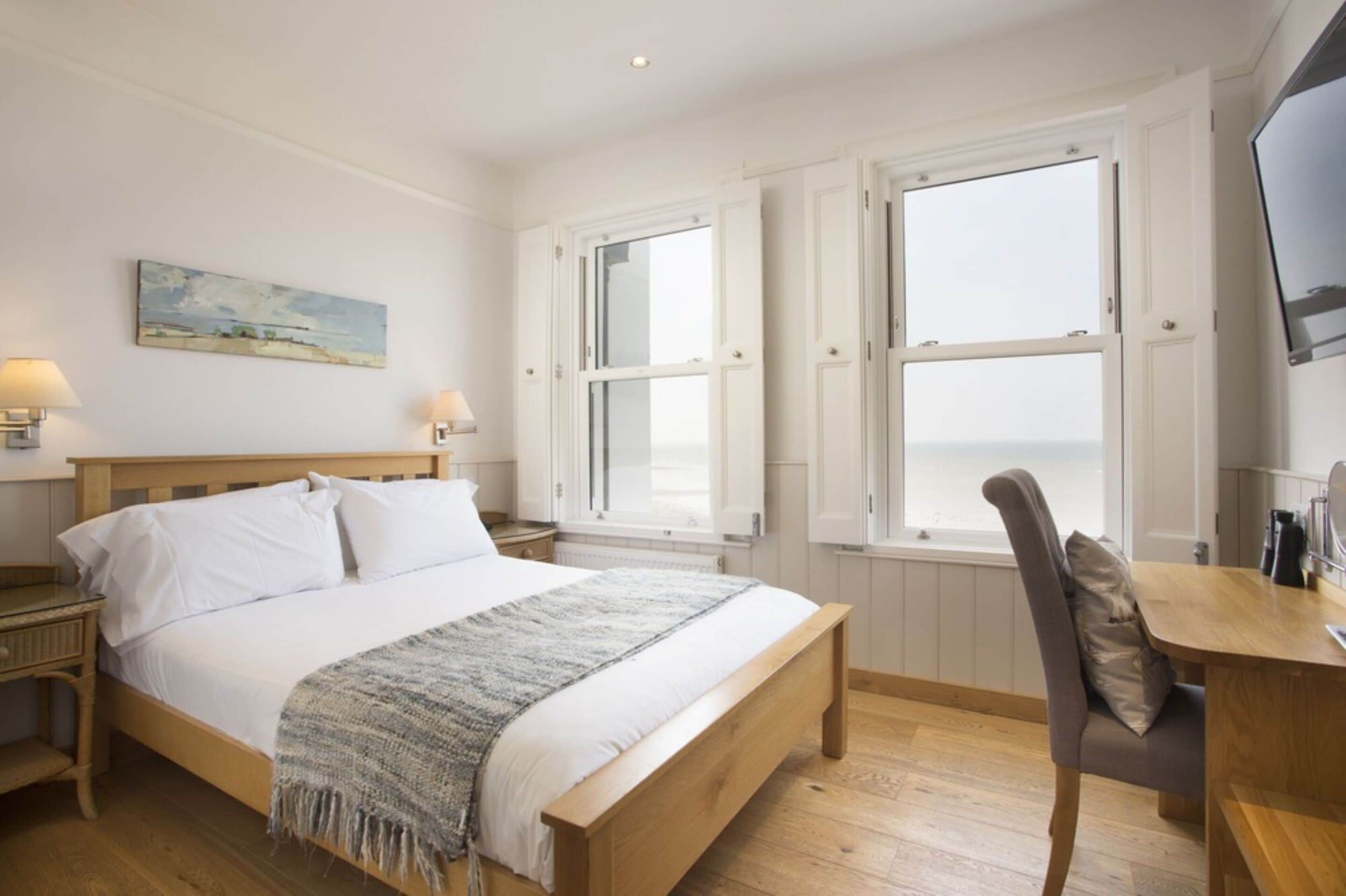 Un dormitorio con una cama de madera, un escritorio y una silla. Hay dos ventanas con marcos blancos y un cuadro en la pared.