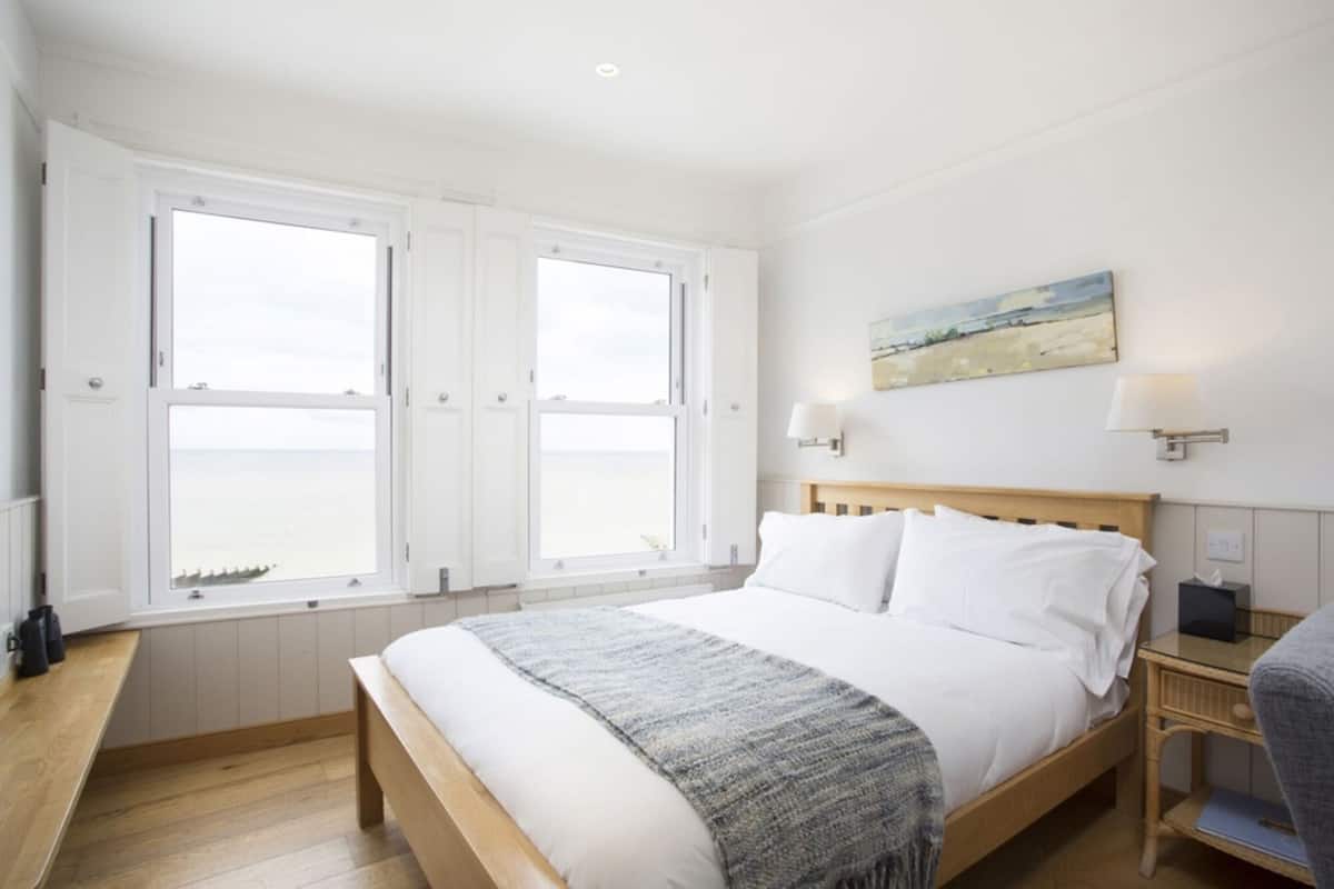 Un dormitorio con una cama grande, dos ventanas, una mesita de noche, una lámpara y un cuadro en la pared.