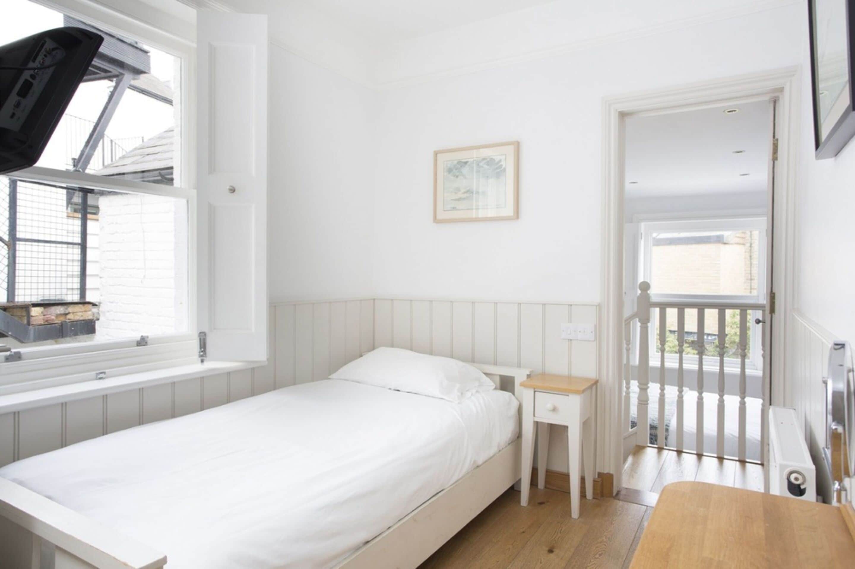 Une chambre avec un lit simple, une petite table de chevet, une fenêtre donnant sur l’extérieur et une porte menant à un balcon.