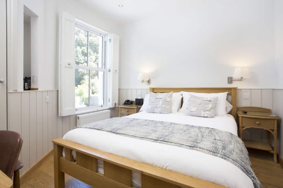 Un dormitorio con una cama de madera, ropa de cama blanca, dos mesitas de noche con lámparas y una ventana con vistas a zonas verdes.