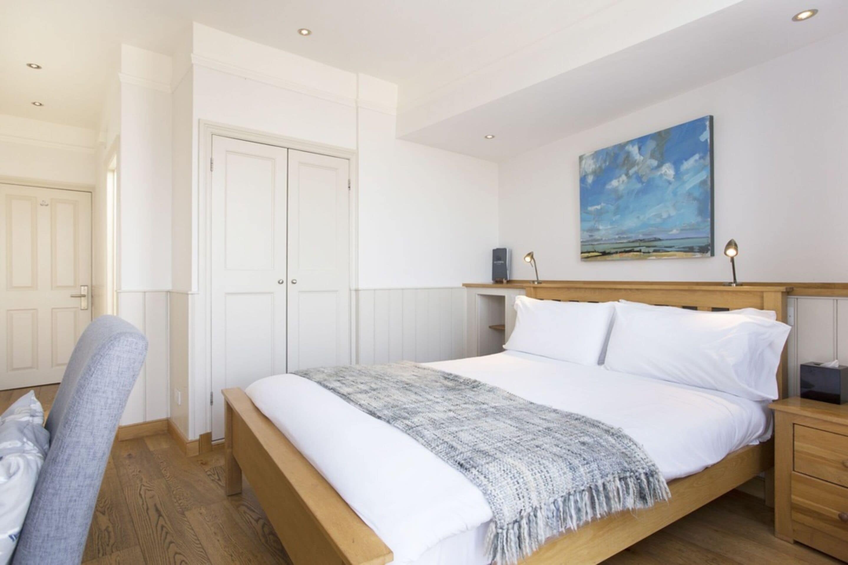 A bedroom with a wooden bed, white bedding, a grey throw blanket, a nightstand, a lamp, and a painting of a beach scene.
