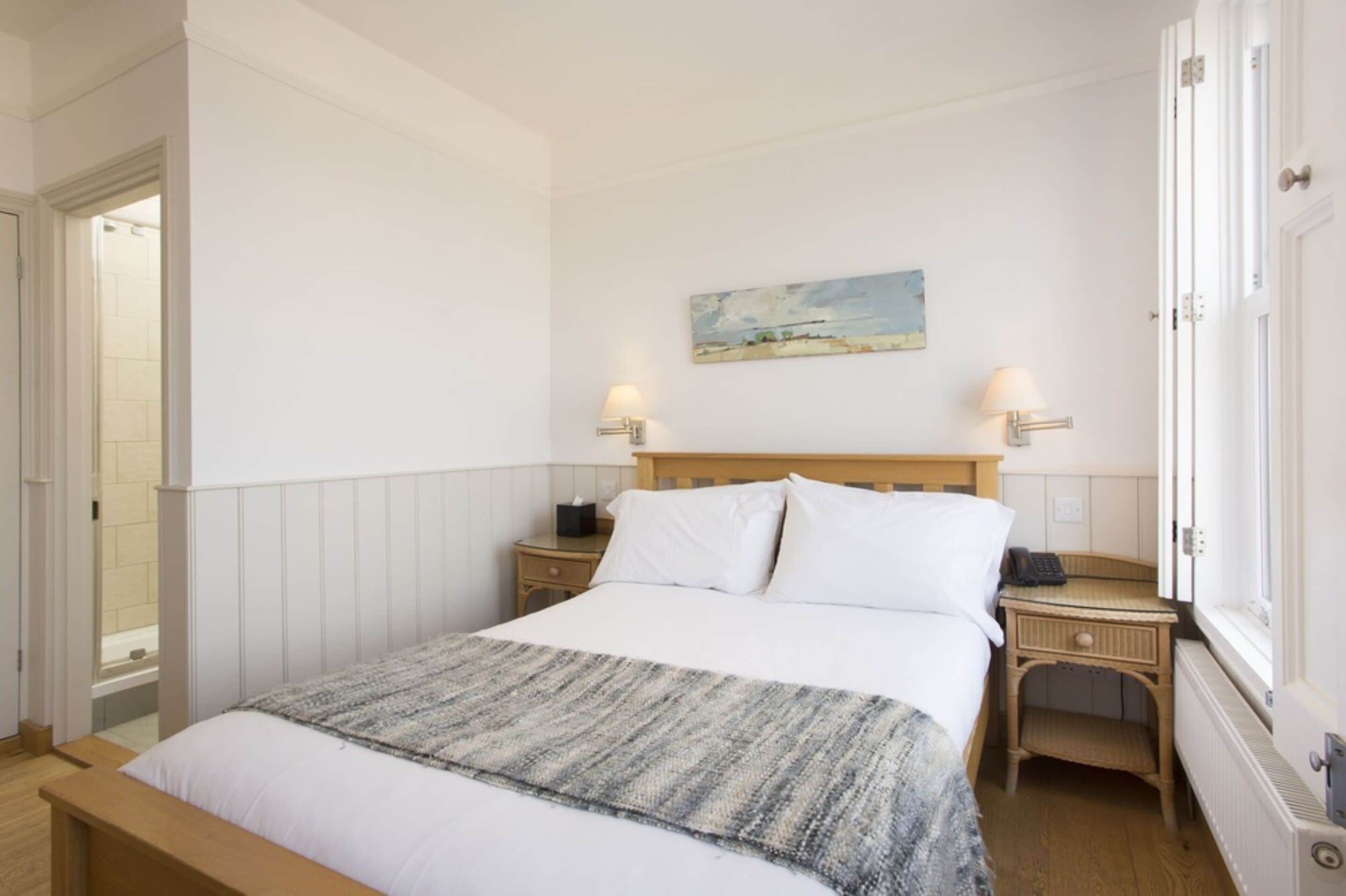 A bedroom with a wooden bed, white bedding, a painting on the wall, two bedside lamps, and a window with a view.