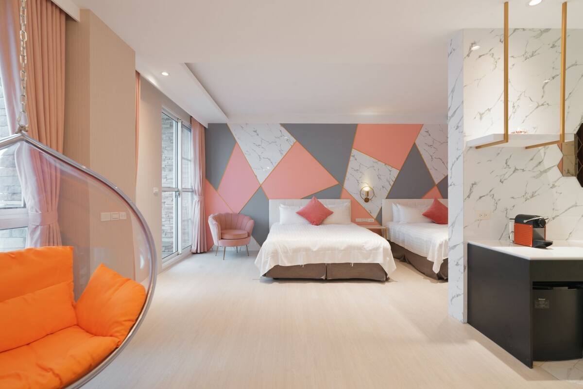 Quarto de hotel moderno com uma janela grande, uma cadeira de vidro curva, duas camas com almofadas cor-de-rosa e uma parede com padrão marmorizado.