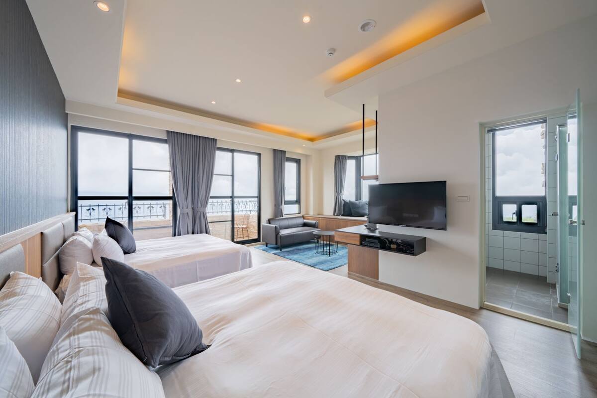 Quarto de hotel moderno com duas camas grandes, uma televisão de tela plana, área de estar com sofá e uma pequena mesa, e varanda com vista.