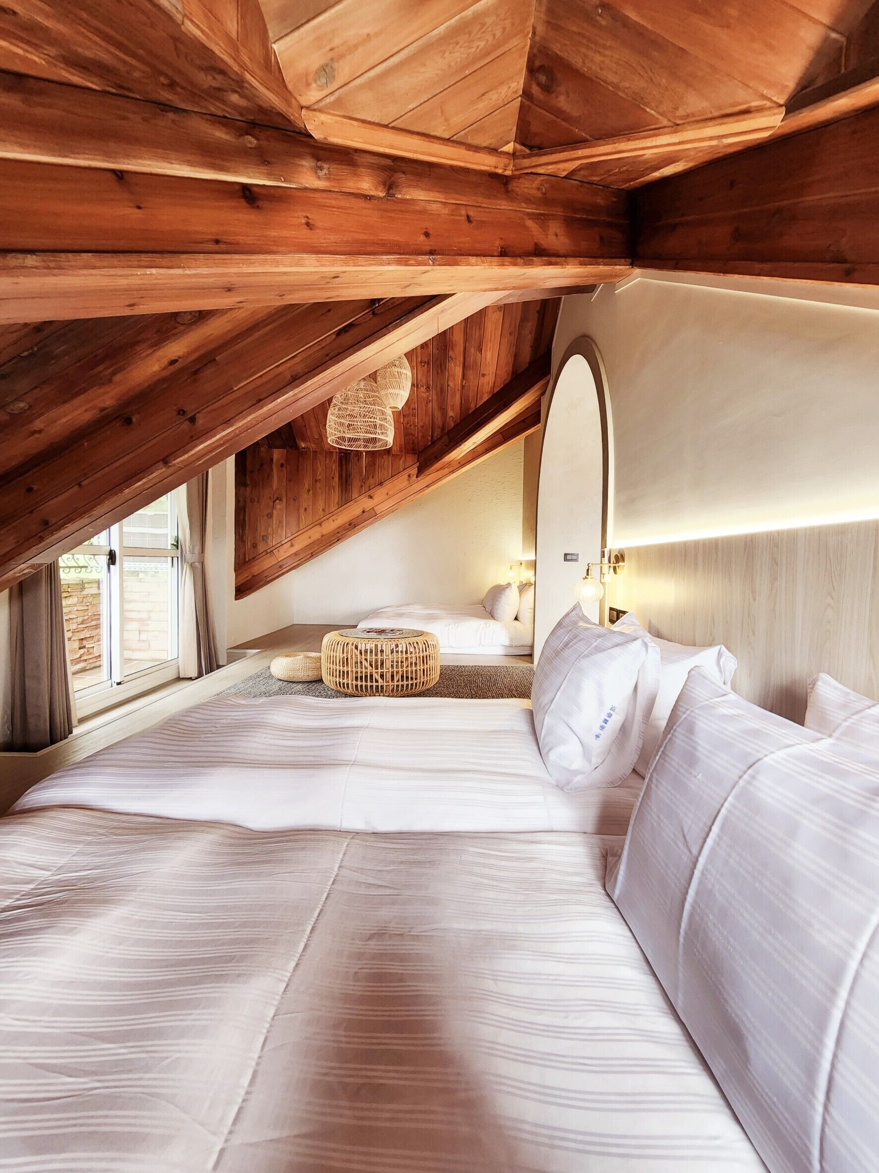 A bedroom with a sloped ceiling, wooden beams, a large bed with white linens, a wicker ottoman, and a bedside lamp.