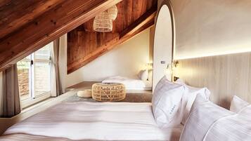 A bedroom with a sloped ceiling, wooden beams, a large bed with white linens, a wicker ottoman, and a bedside lamp.