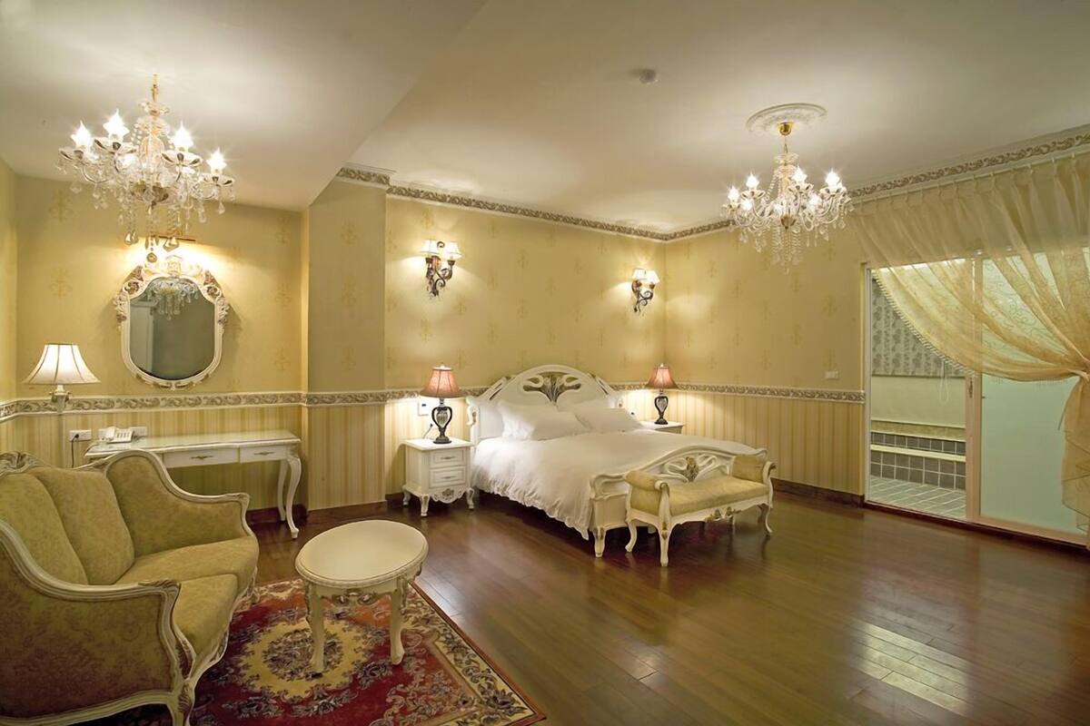 Quarto clássico e elegantemente decorado, com uma cama grande, duas mesas de cabeceira, um espaço de estar com sofá e poltrona, e um lustre.
