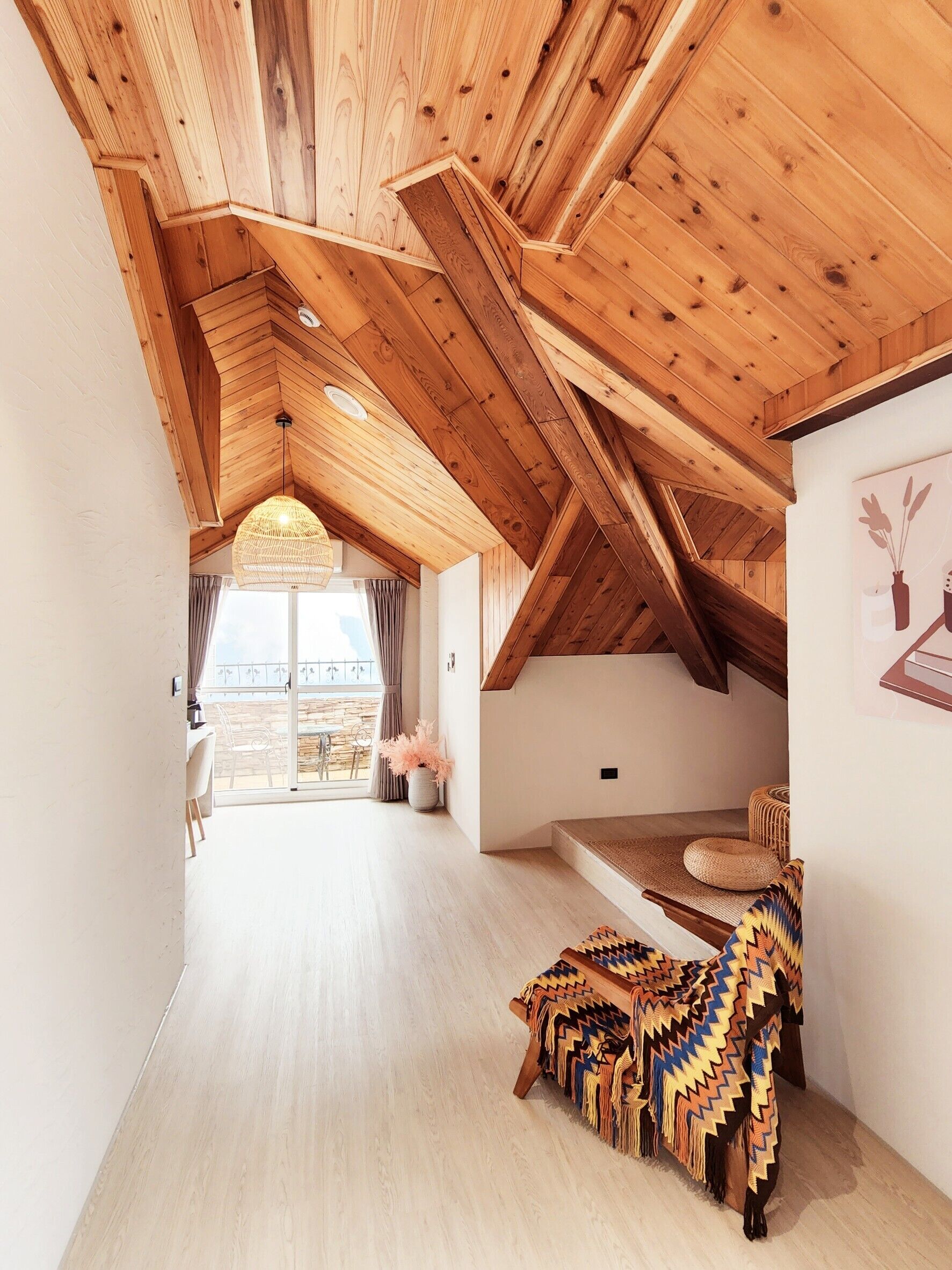 A spacious loft bedroom with a high ceiling, wooden beams, a large window, and a cozy seating area with a patterned blanket.