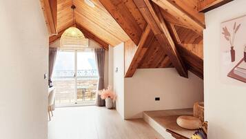 A spacious loft bedroom with a high ceiling, wooden beams, a large window, and a cozy seating area with a patterned blanket.