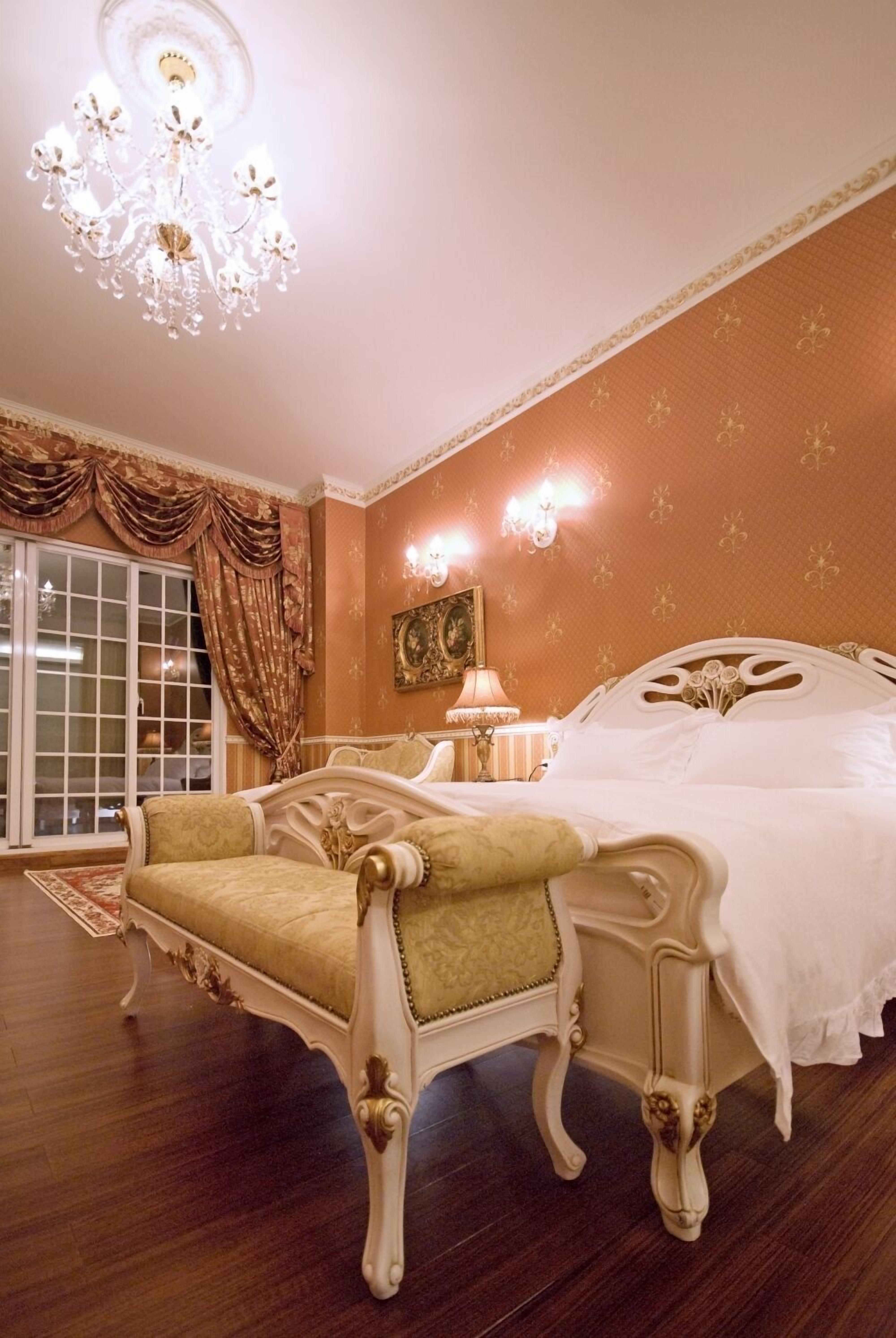 A bedroom with a large bed, ornate chair, chandelier, and a large window with curtains.