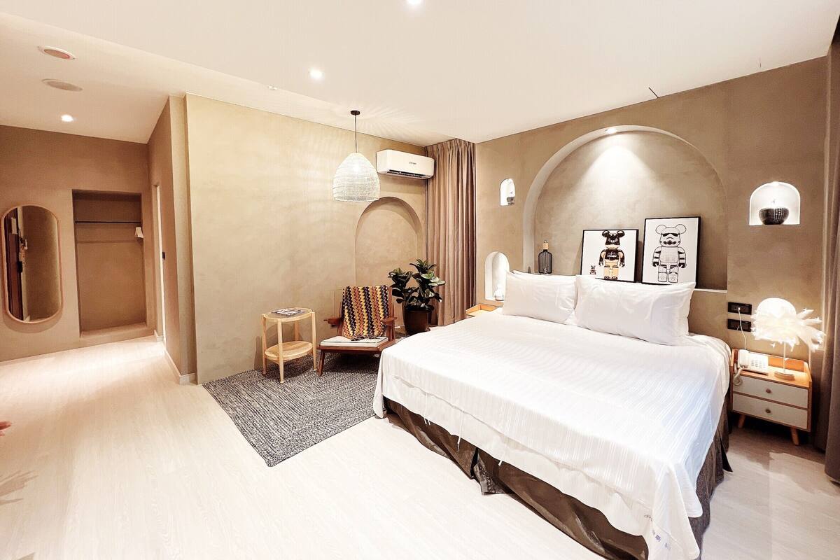 Quarto moderno com uma cama grande, uma mesa de cabeceira com abajur, uma mesinha lateral e um vaso com planta.