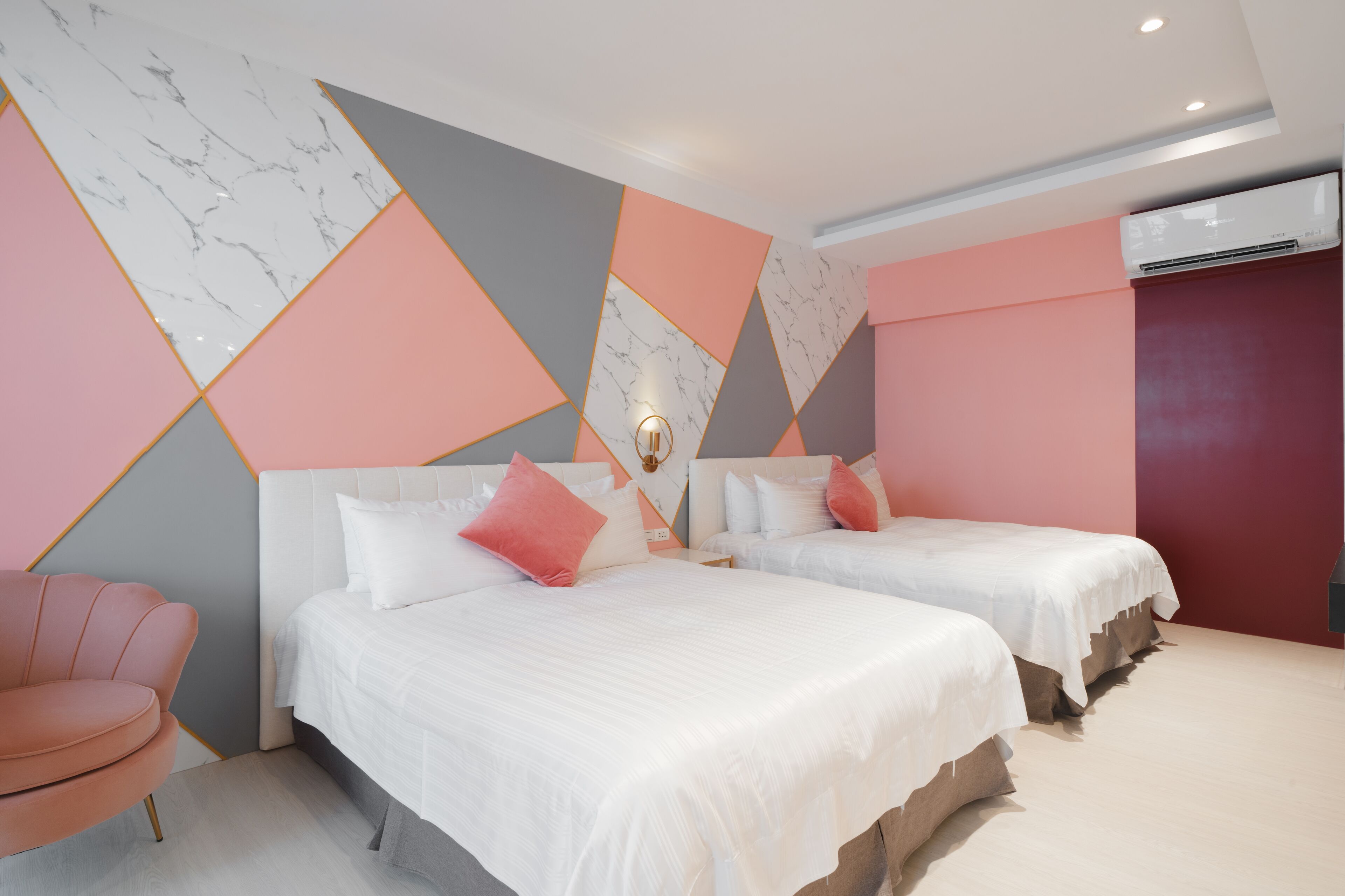 Habitación de hotel con dos camas, una silla rosa y una pared con diseño geométrico.