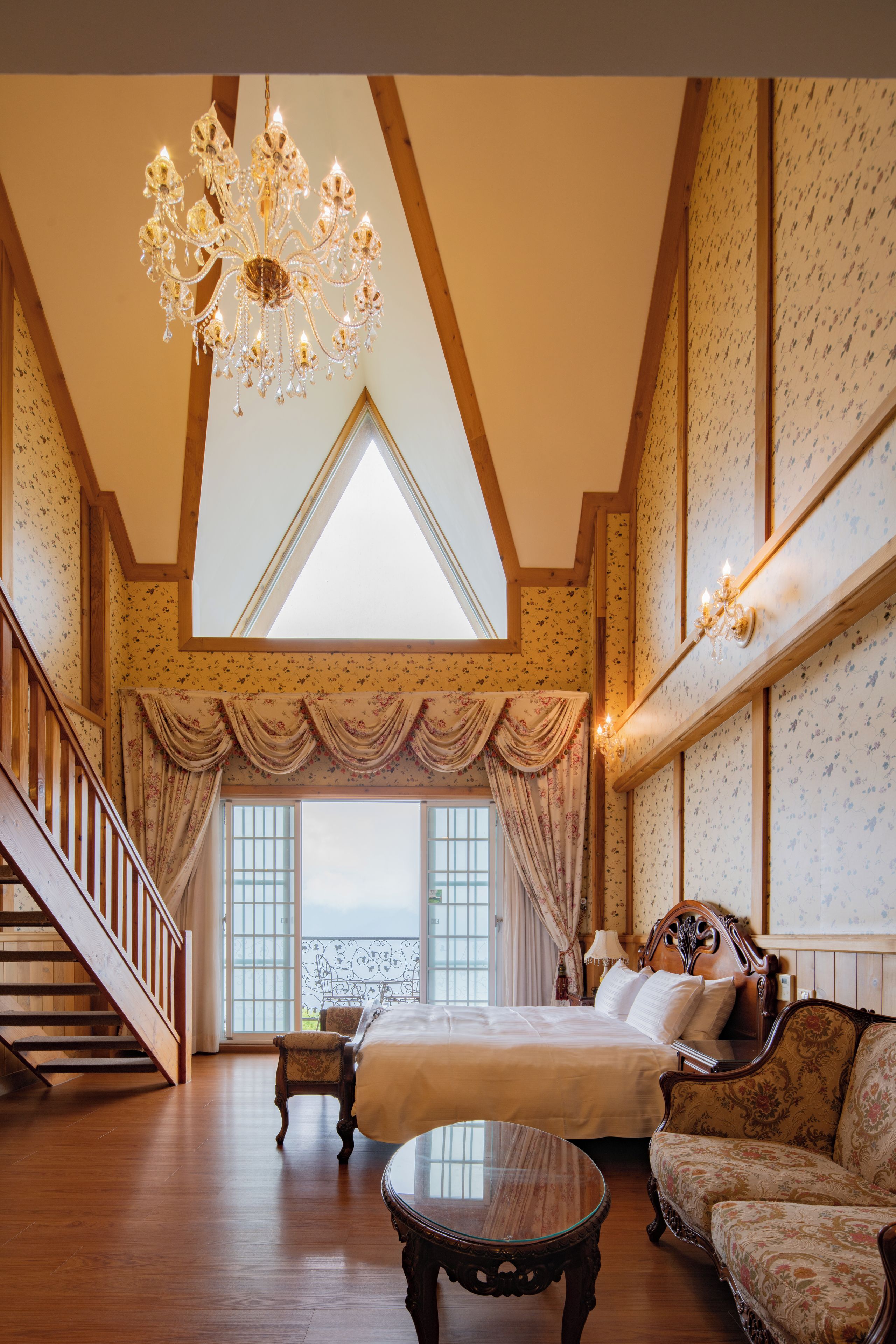 A spacious bedroom with a large bed, a sitting area with a sofa, a chandelier, and a staircase leading to an upper level.
