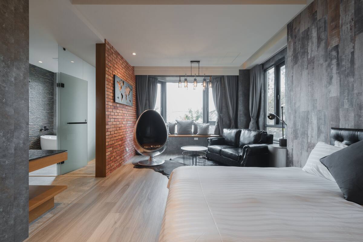 Quarto de hotel moderno com uma cama grande, área de estar com um sofá de couro preto, uma cadeira redonda preta, uma mesa redonda pequena e uma parede decorada com tijolos.