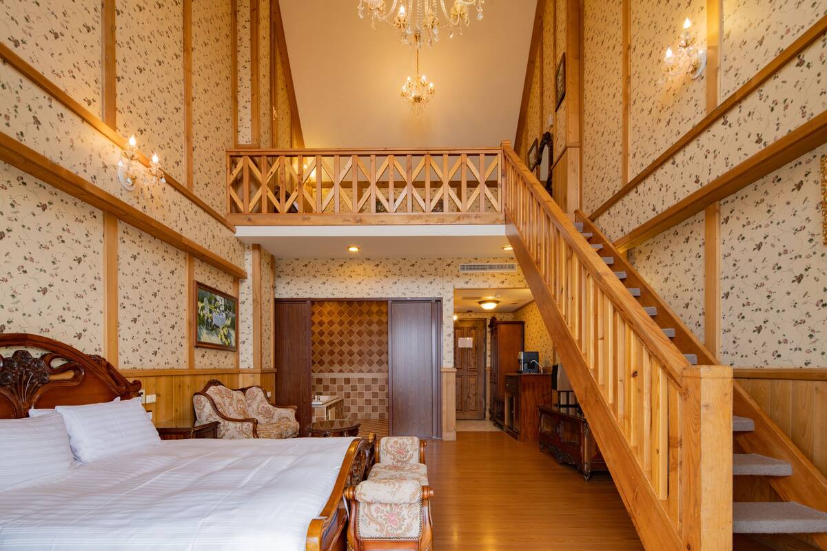 Quarto espaçoso com uma cama grande, escada de madeira e um pequeno espaço de estar com cadeiras.