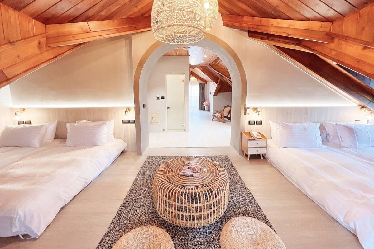 Quarto com pé-direito alto, vigas de madeira, duas camas, um banquinho de fibra central e um corredor que dá para outro quarto.