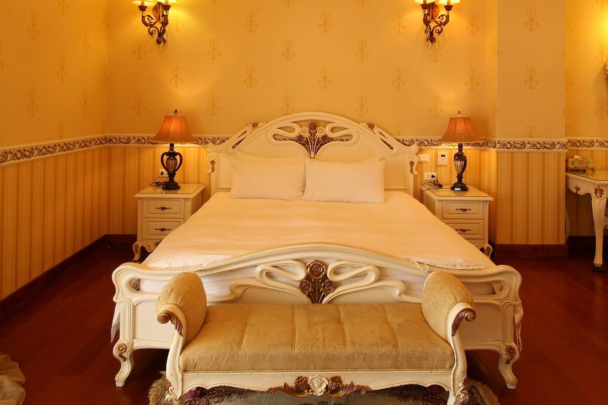 Quarto com cama grande, lustre ornamentado, duas mesas de cabeceira com abajures e tapete estampado.