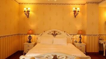 A bedroom with a large bed, ornate chandelier, two bedside tables with lamps, and a patterned rug.