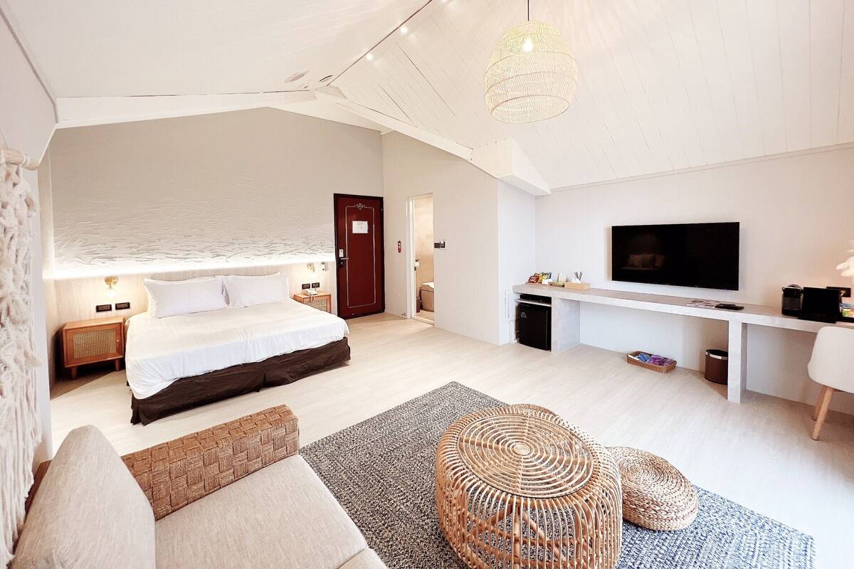Quarto moderno com cama, mesa de cabeceira, luminária e televisão sobre um suporte.