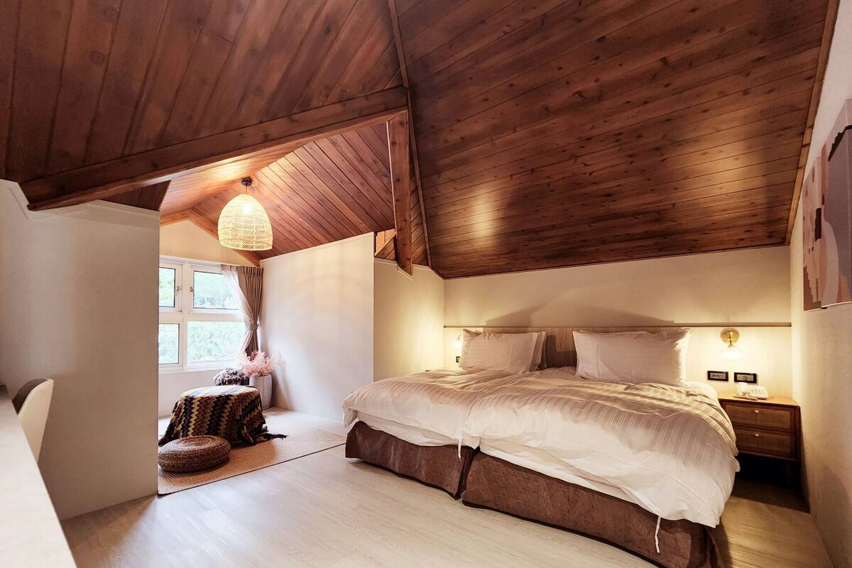 Um quarto acolhedor com teto de madeira, uma cama grande com roupa de cama branca, uma mesa de cabeceira com abajur e uma janela com cortinas.