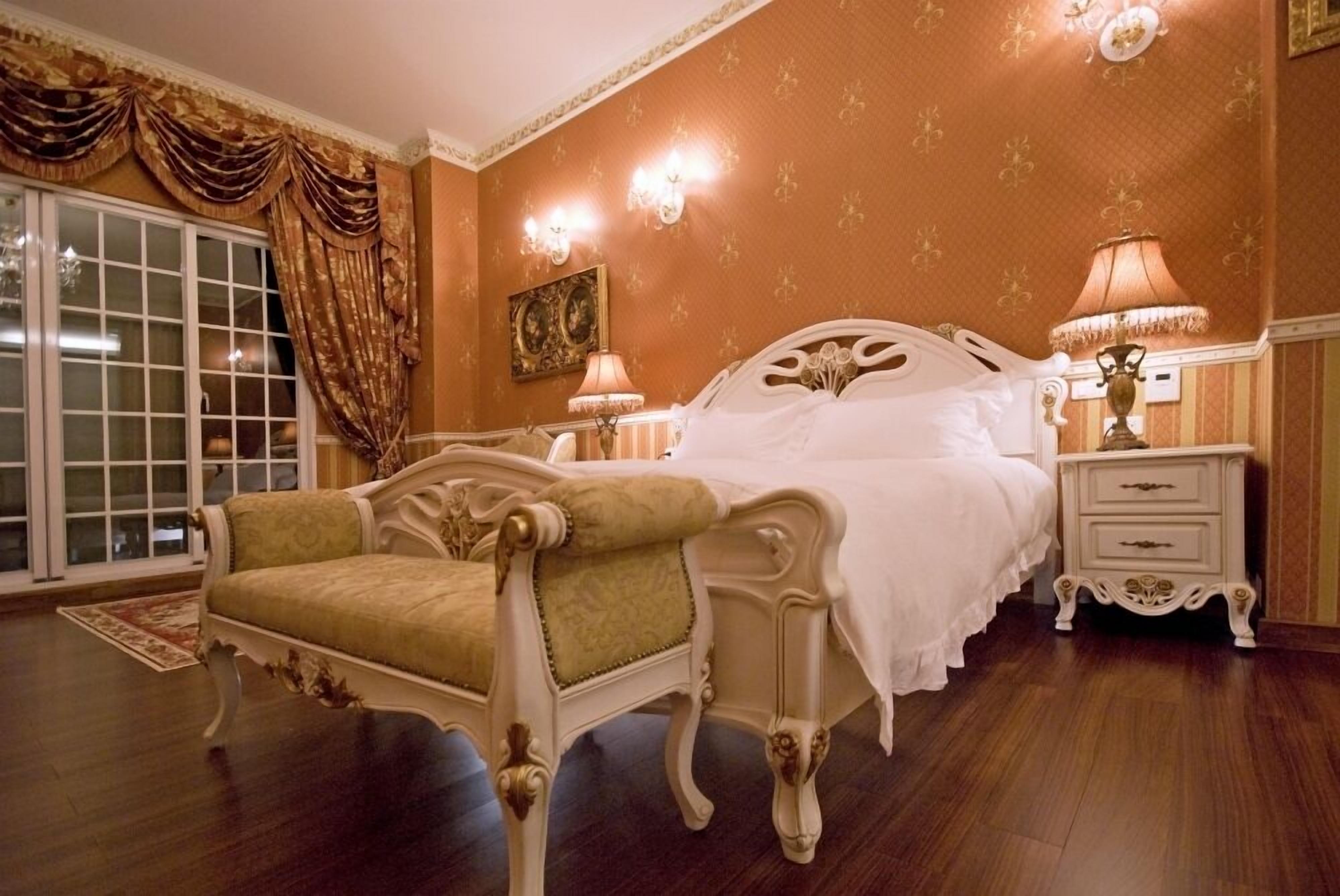 A classic bedroom with a large bed, a chaise lounge, a nightstand, and a wall-mounted lamp.