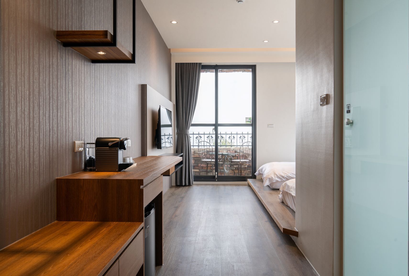 A modern hotel room with a wooden desk, a flat-screen TV, a balcony with a view, and a bed.
