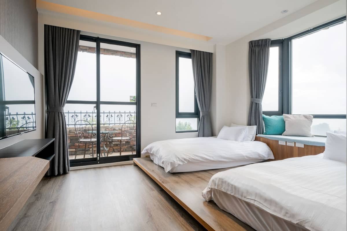 Quarto de hotel moderno com duas camas, televisão, varanda com área de estar e janelas amplas.