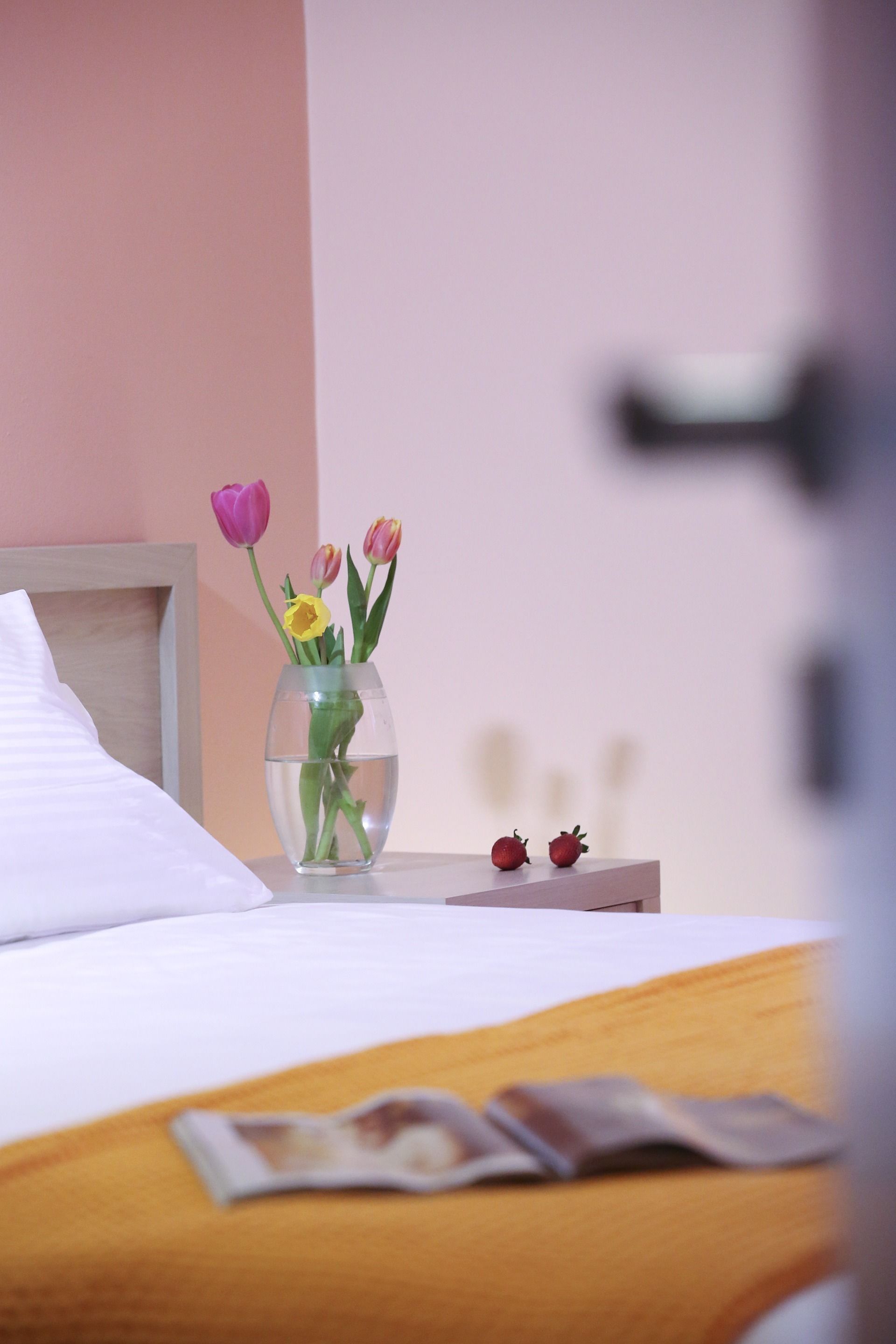 A bed with white bedding, a clear vase with flowers, and a wooden nightstand.