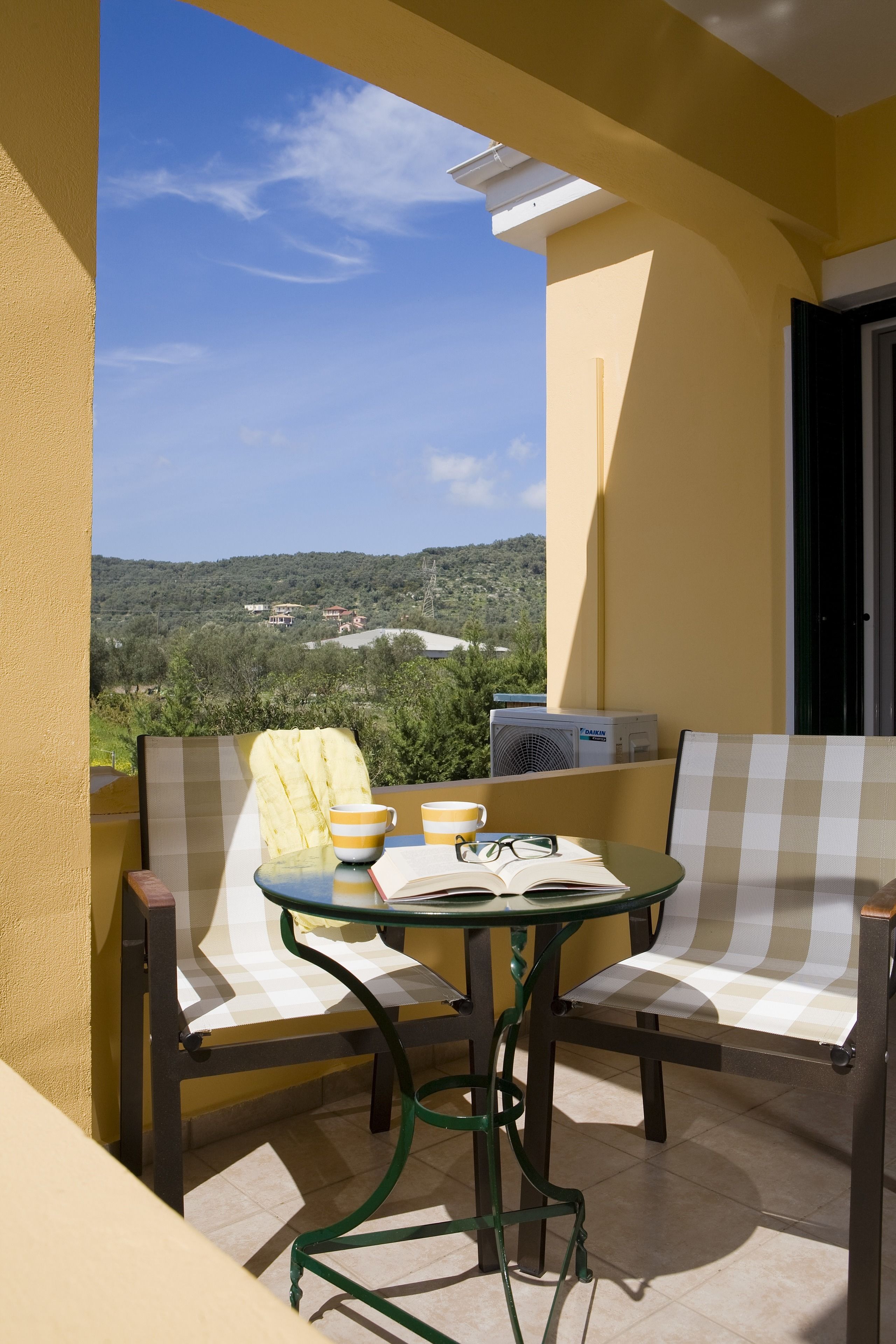 A balcony with a checkered sofa, a small table with a book and a cup, and a view of a green landscape.