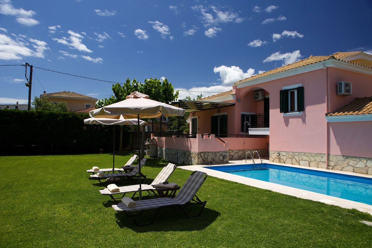 Un área junto a la piscina con sillones y sombrillas, piscina y un edificio rosa con tejado de azulejos.