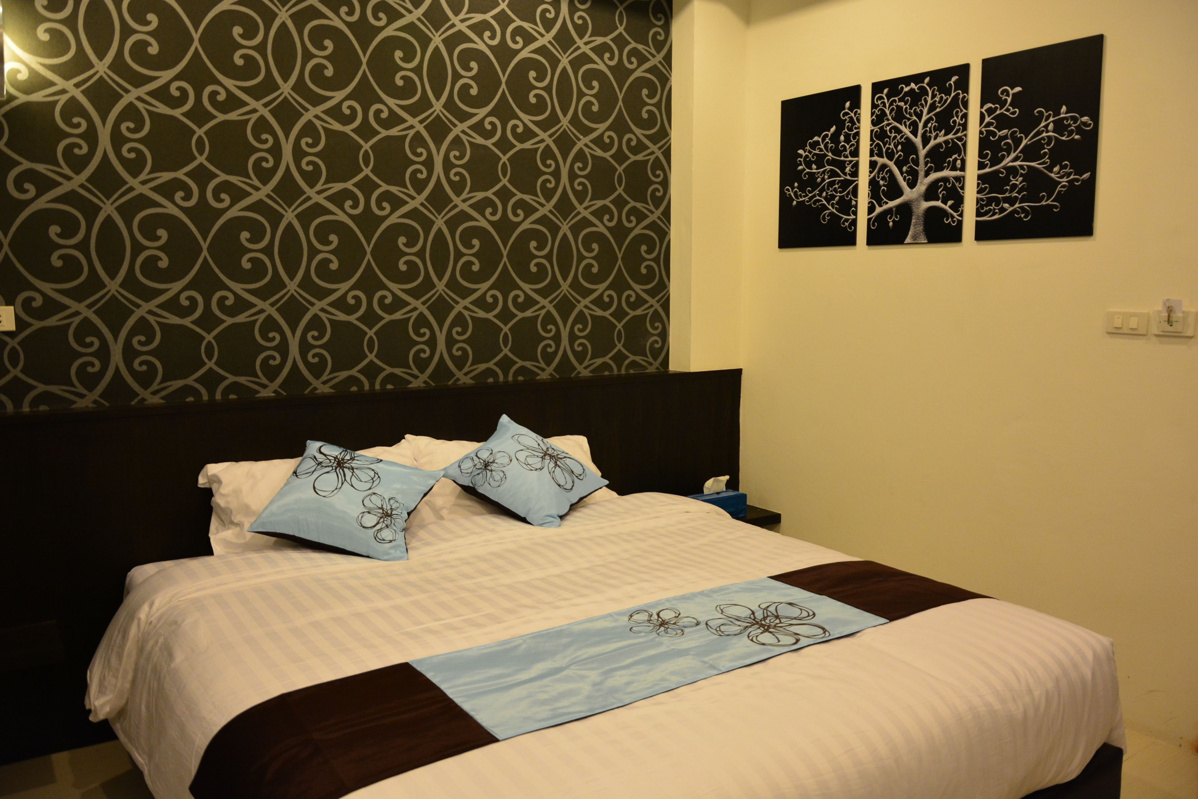 A neatly made bed with a patterned headboard, a blue decorative runner, and two wall-mounted paintings.