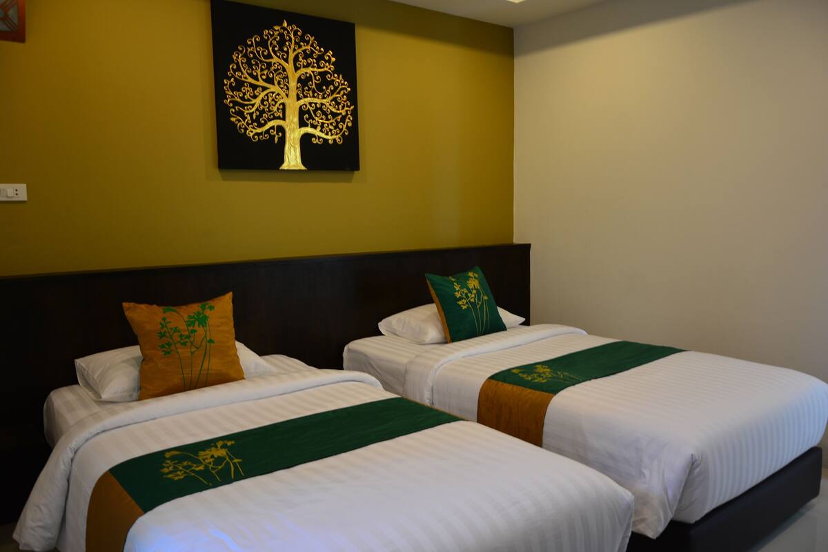 A hotel room with two beds, each with white linens and green and orange decorative pillows. A large tree artwork is displayed on the wall above the beds.