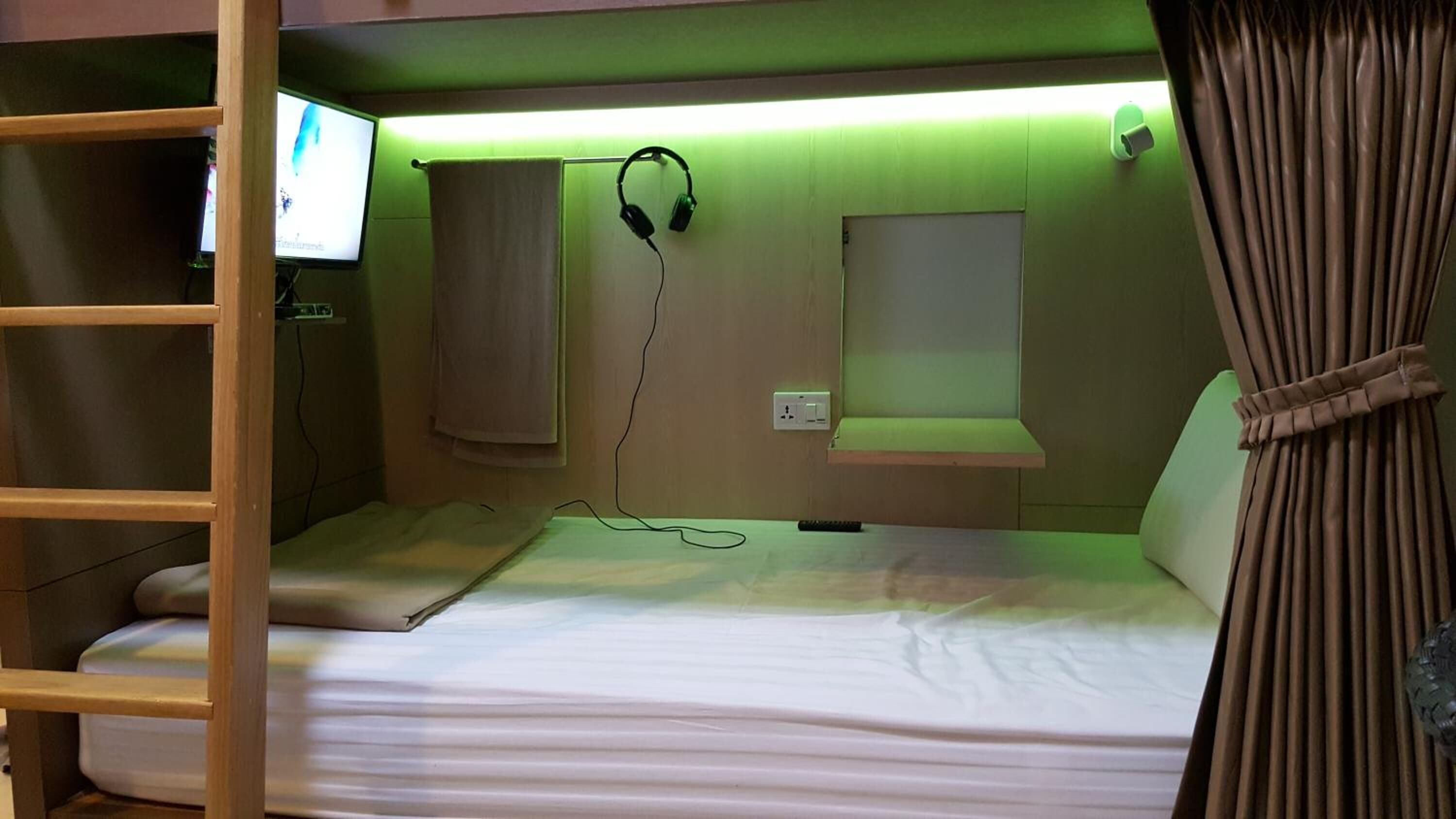 A bunk bed with a television mounted on the wall, headphones hanging on the wall, and a small window with curtains.