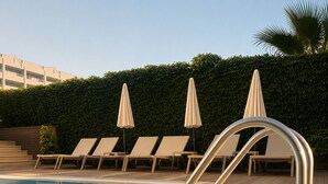 Piscina com espreguiçadeiras e guarda-sóis, cercada por um viveiro e um edifício ao fundo.
