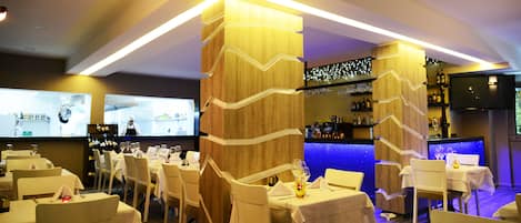 A restaurant with multiple tables and chairs, a bar area, and a television screen displaying a chef.