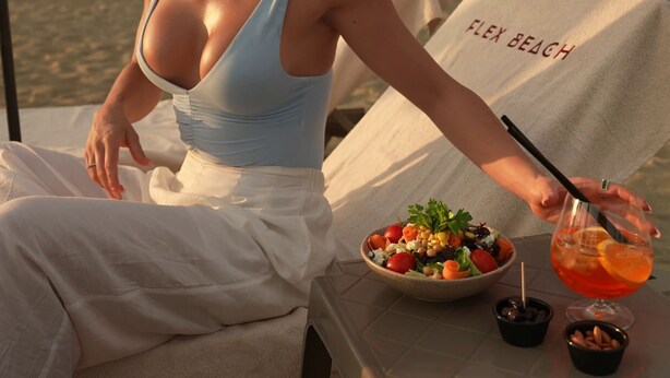 Uma pessoa sentada à mesa com uma salada e uma bebida, com um guarda-sol de praia identificado como 