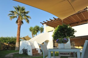 A patio area with a covered table and chairs, a palm tree, and a white building with steps leading up to a door.