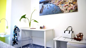 A room with a white desk, a potted plant, a painting of a coastal town, and a bed with a patterned bedspread. - Il Leone di Mare (Procida)