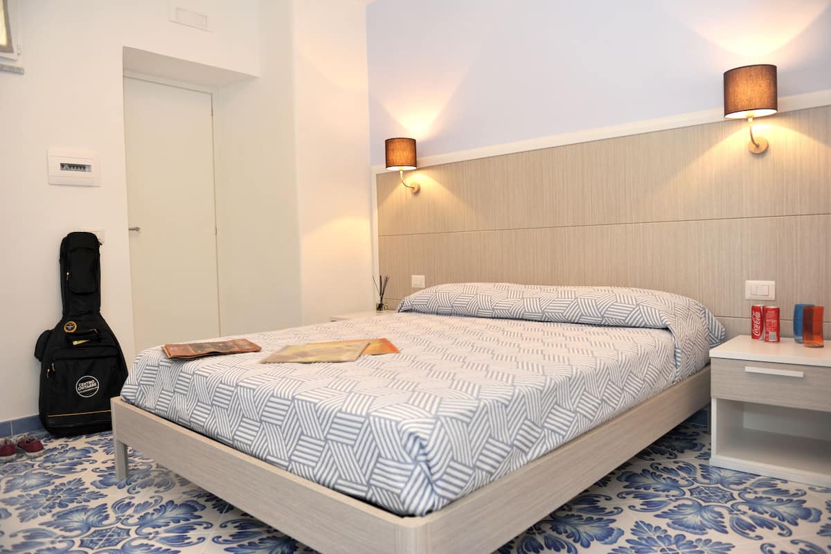 A bedroom with a bed, a nightstand, a lamp, a guitar, and a door.