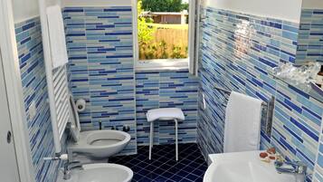 Een badkamer met toilet, bidet en wastafel, betegeld in blauw en wit, met een raam dat uitzicht biedt op groen buiten.