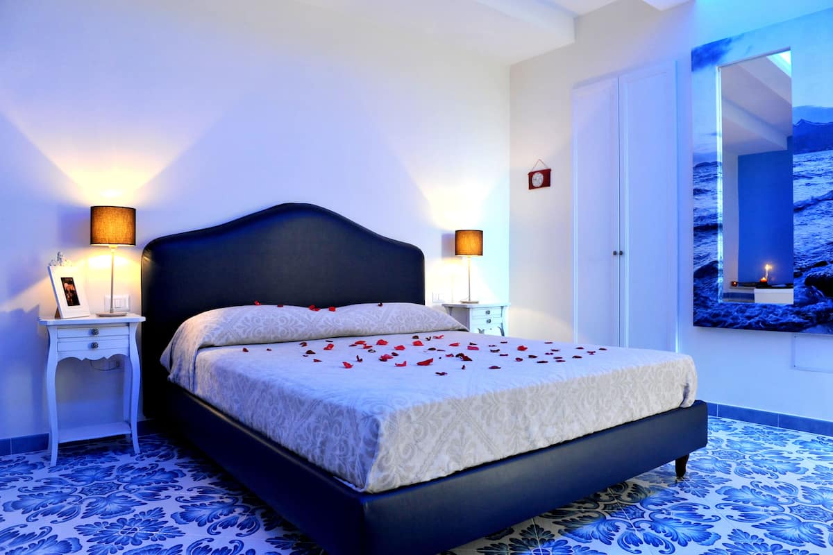 A bedroom with a large bed, two bedside tables with lamps, a nightstand with a picture frame, and a wall-mounted TV displaying a beach scene.