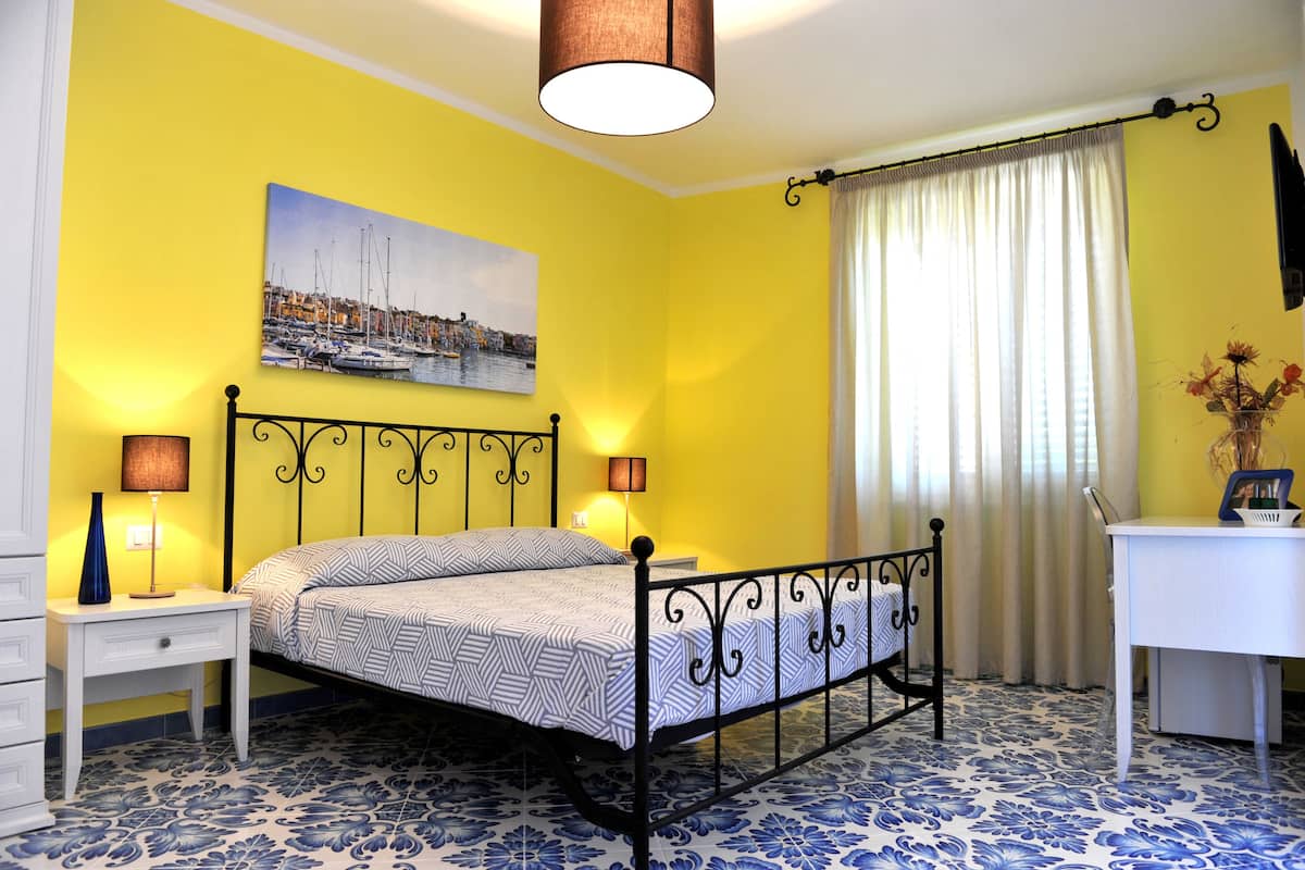 A bedroom with a bed, bedside tables, a desk, a chair, a lamp, a painting, and a curtain.