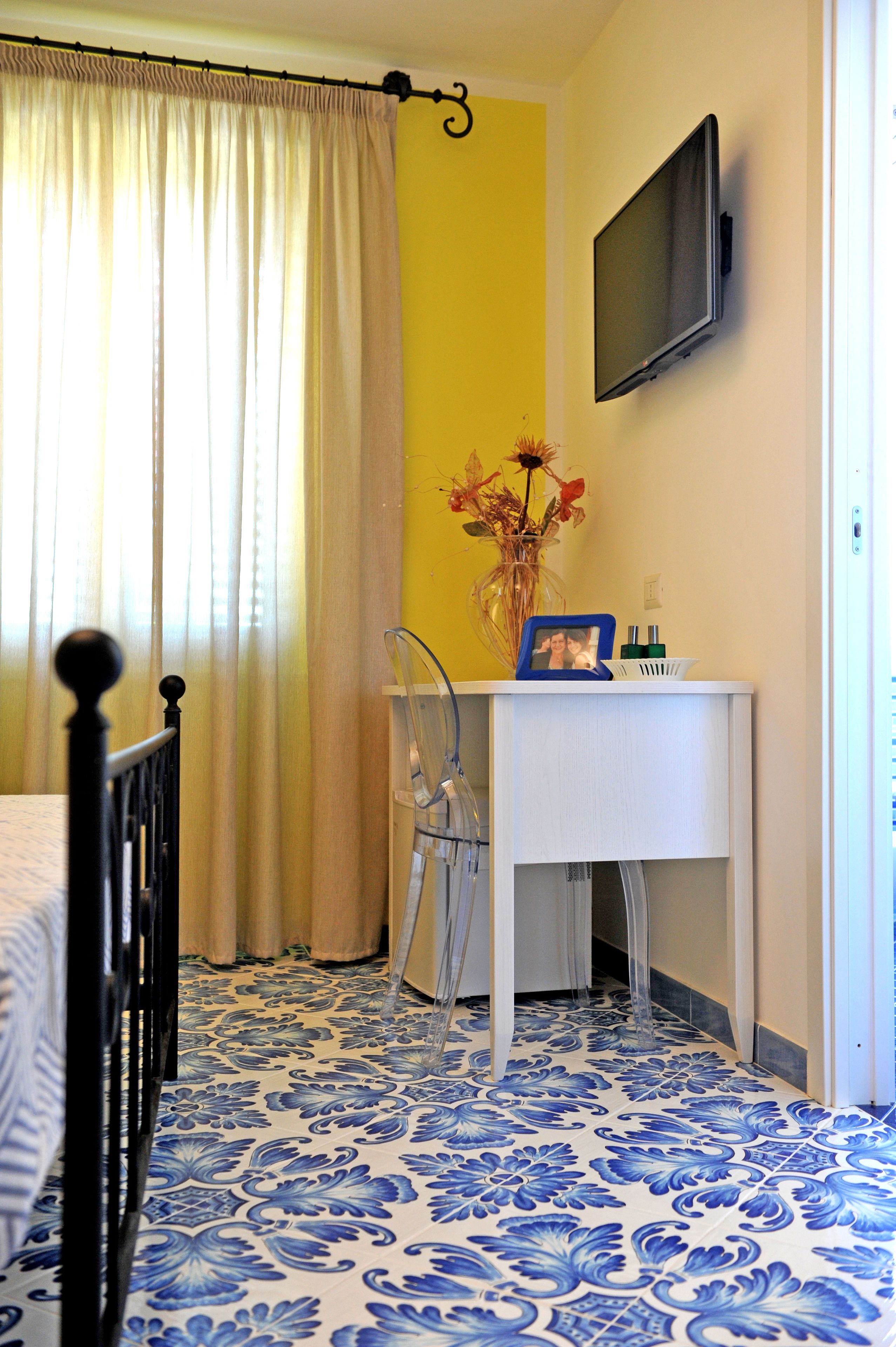 A bedroom with a bed, a desk with a chair, a TV mounted on the wall, a vase with flowers, and a yellow wall.