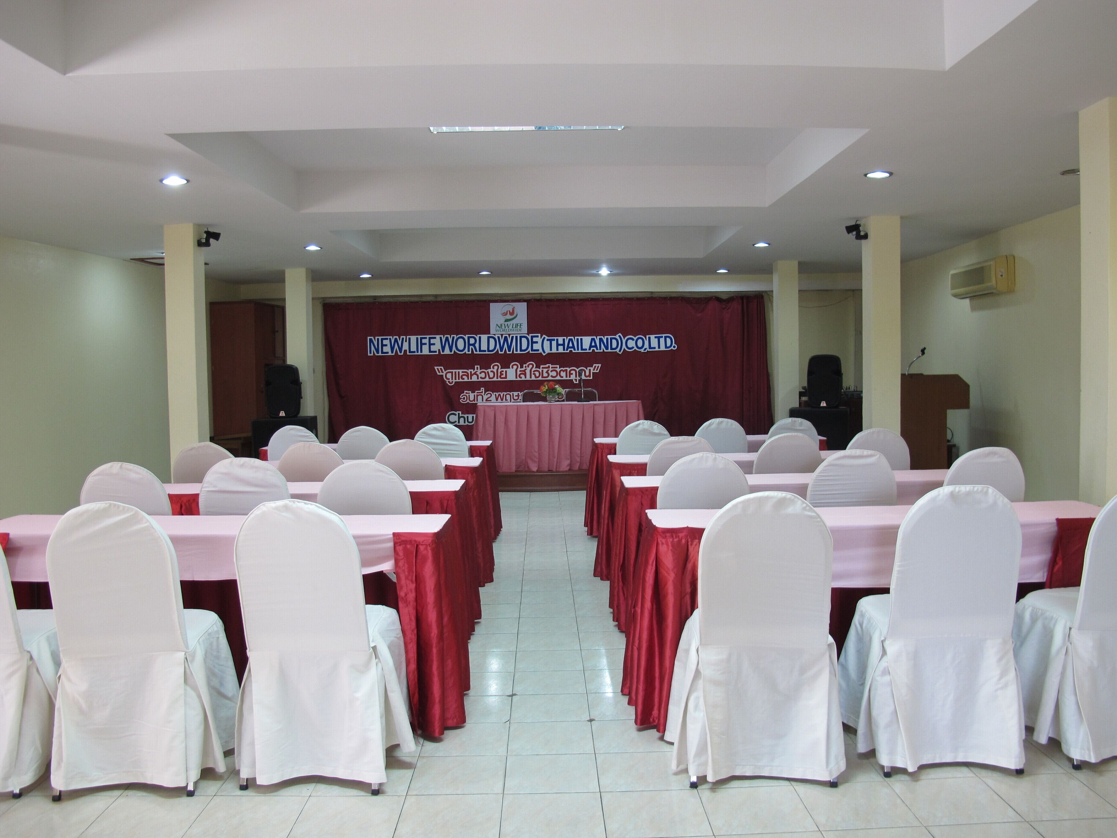 Un salón de conferencias con filas de mesas y sillas, un atril y un cartel que dice "NEW LIFE WORLDWIDE (THAILAND) CO., LTD.".