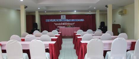 Un salón de conferencias con filas de mesas y sillas, un atril y un cartel que dice "NEW LIFE WORLDWIDE (THAILAND) CO., LTD.".