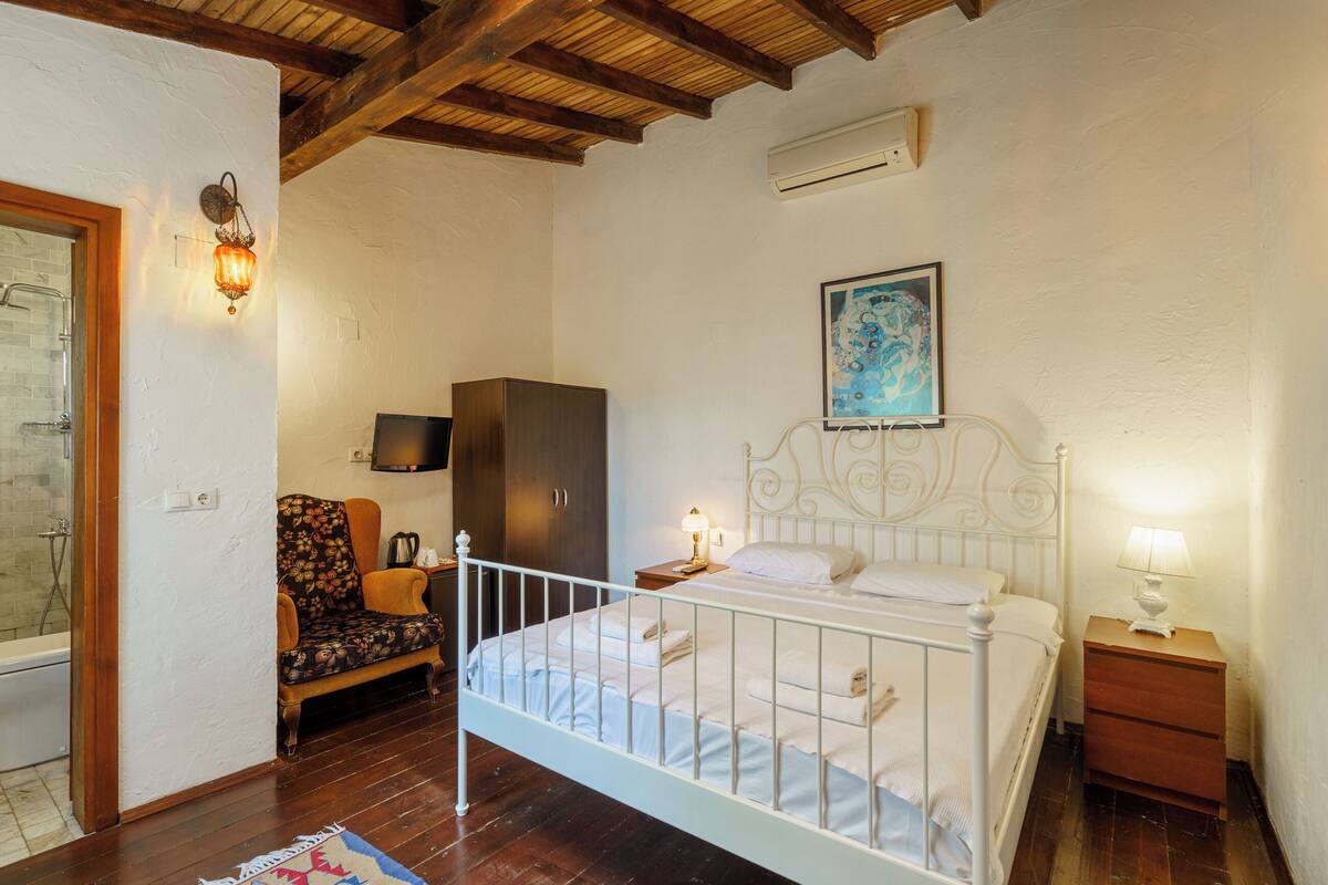 A bedroom with a white metal bed, a wooden wardrobe, a chair, a bedside table with a lamp, and a painting on the wall.