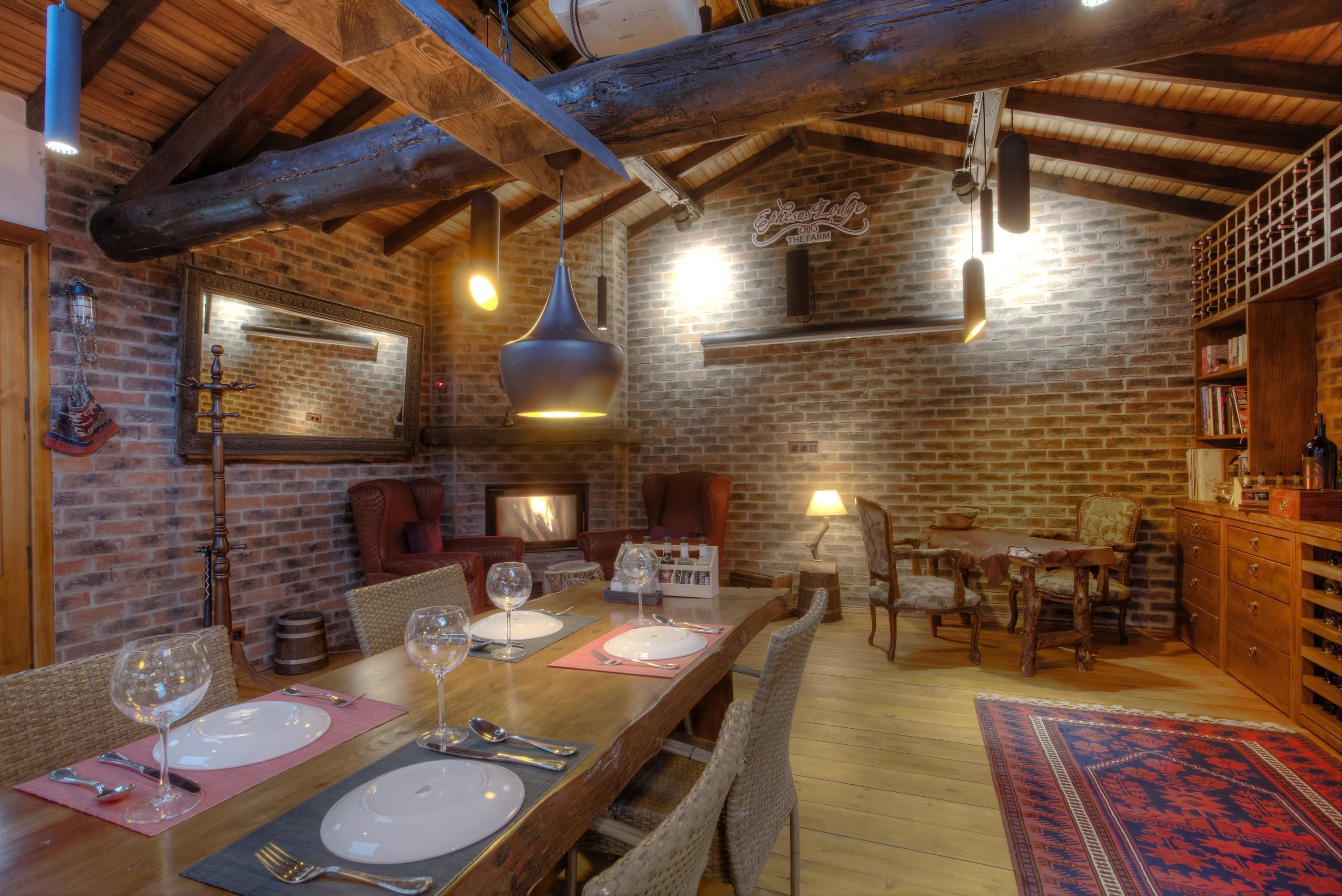 A rustic dining area with a long wooden table set for four, surrounded by wicker chairs. A brick wall and exposed wooden beams are visible in the background.