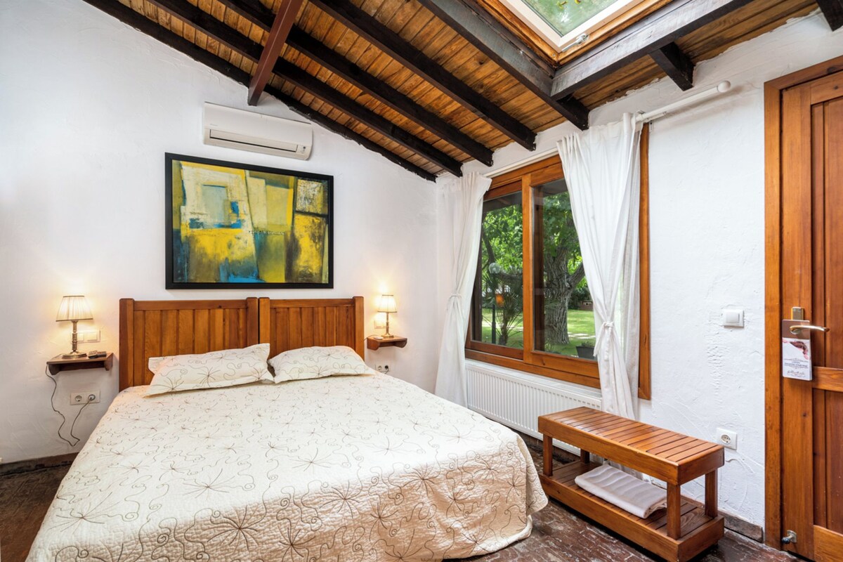A bedroom with a wooden headboard, a large bed with a patterned bedspread, a painting above the bed, a window with curtains, and a small wooden bench by the window.