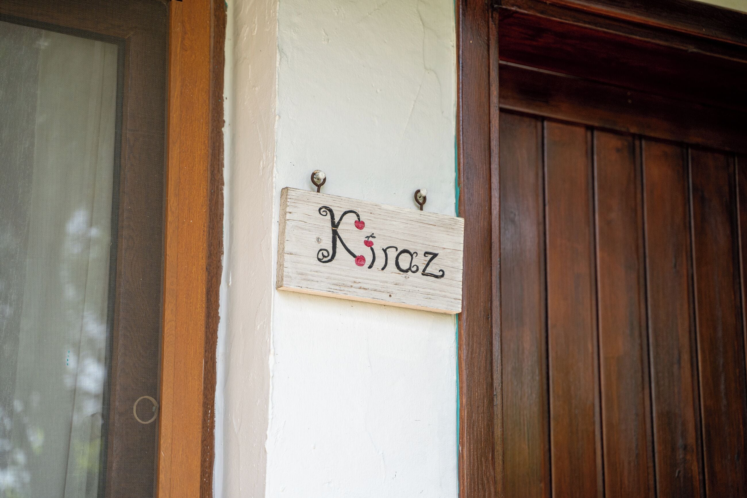 A wooden sign with the name 'Kira' on a white wall, with a wooden door and a window with a mesh screen to the left.